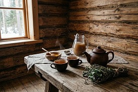 Глиняный чайник, чашки травяного чая, мёд, кувшин с напитком и пучок трав на столе в бане